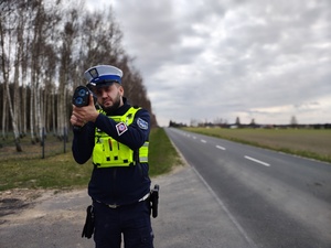 Policjant stoi przy drodze, mierzy urządzeniem prędkość przejeżdżających samochodów.