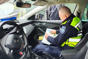 Policjant siedzi w radiowozie, wypisuje mandat, przy drzwiach samochodu stoi osoba ubrana po cywilnemu.