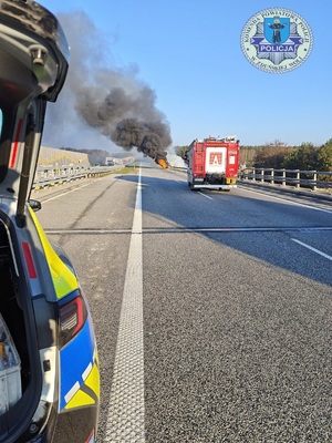 Droga, w oddali czarny dym palącego się auta, za pojazdem stoi samochód strażacki, a bliżej radiowóz.