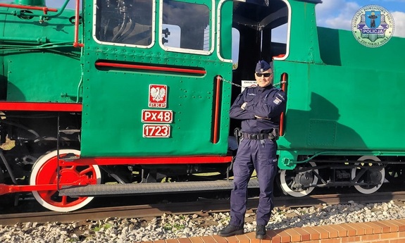 Policjant stoi pod muzealną lokomotywą. Ma założone okulary słoneczne. Ręce ma splecione na ramionach.