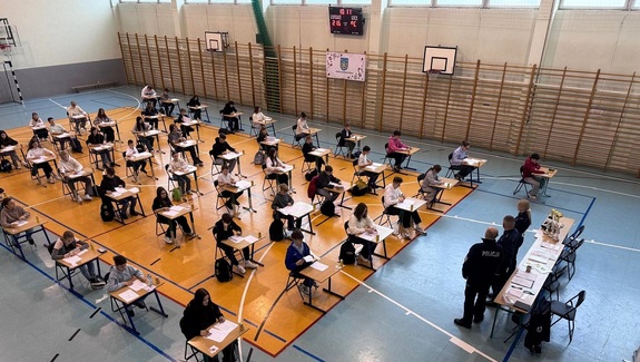 Zdjęcie z góry, pokazujące salę gimnastyczną. Są poustawiane krzesełka i stoliki, przy których uczniowie piszą test.