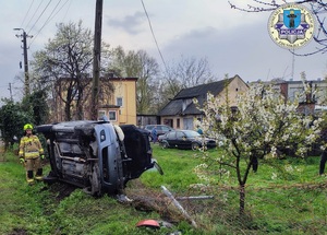Samochód leży na boku, na trawie, na terenie posesji, uszkodzony płot, obok stoi strażak.