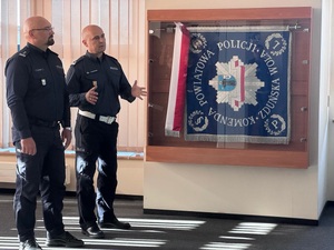 Dwaj policjanci stoją obok siebie, mówią do kogoś przed nimi. Na ścianie obok nich wisi sztandar jednostki.