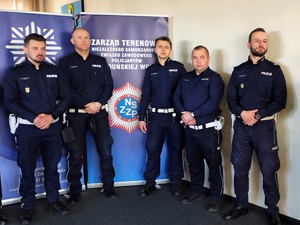 Pod ścianą, w rzędzie stoją policjanci.