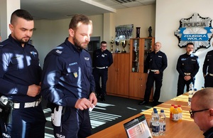 Policjanci stoją w kolejce do losowania swoich miejsc. Policjant pokazuje komisji karteczkę ze swoim wylosowanym numerkiem.