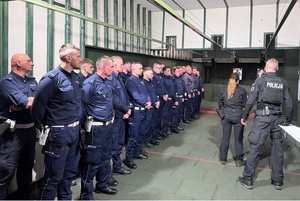 Policjanci stoją w rzędzie pod ścianą na strzelnicy. Przed nimi stoi instruktorka oraz instruktor strzelania.