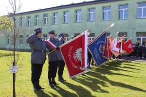 Poczet sztandarowy Policji, policjanci salutują, za nimi stoją kolejne poczty, w tle budynek szkoły.