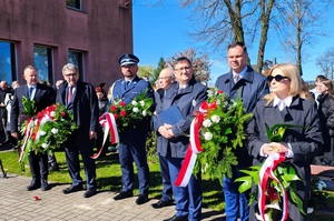 Goście uroczystości stoją z kwiatami, które złożą przez tablicą pamiątkową.