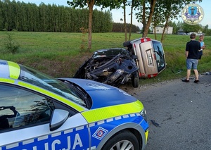 Na poboczu drogi stoi radiowóz oraz dwa rozbite samochody, jeden z nich leży przewrócony na prawym boku. Obok samochodów stoją dwie osoby.