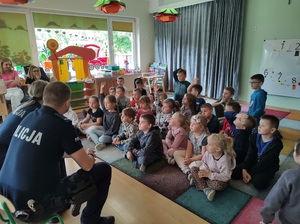 Dzieci siedzą na dywanie. Przed nimi siedzą na krzesełkach policjanci, mówią do dzieci.