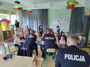 Dzieci siedzą na dywanie, przed nimi na krzesełkach siedzą policjanci, mówią do dzieci.