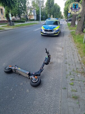 Na jezdni leży przewrócona hulajnoga, za nią stoi policyjny radiowóz.