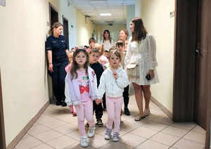 Dzieci idą w parach korytarzem, obok opiekunki i policjantka.