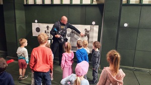 Dzieci słuchają co mówi do nich policjant, który pokazuje im na ścianie powieszone różnego rodzaju tarcze do strzelania.
