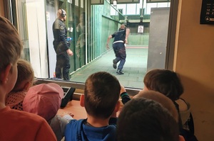 Dzieci przez szybę oglądają ćwiczenia policjanta na strzelnicy.