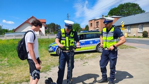 Dwaj policjanci rozmawiają z chłopakiem, którzy ma hulajnogę. Teren miasta, widać w tle zabudowania.