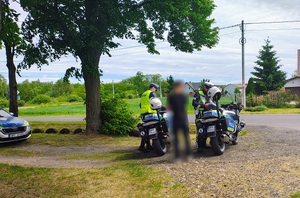 Policjanci stoją przy motorach, między nimi stoi osoba. W tle droga.