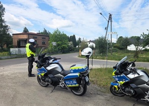 Policjant patrzy przez urządzenie z jaką prędkością jeżdżą pojazdy, obok stoją dwa służbowe motory.
