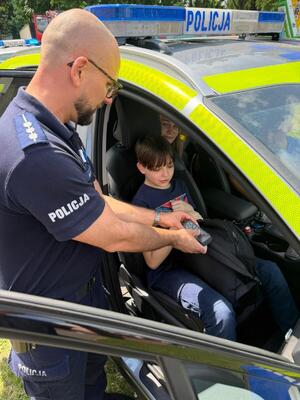 Policjant stoi przy radiowozie. Pokazuje siedzącym w środku dzieciom wnętrze pojazdu.