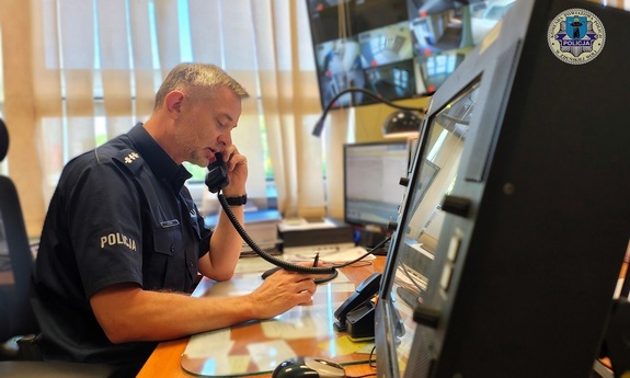 Policjant siedzi za biurkiem, rozmawia przez telefon, przed nim stoją monitory.