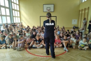 W sali gimnastycznej, na podłodze siedzą uczniowie. Na wprost nich stoi policjantka, mówi do uczniów.