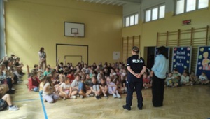 W sali gimnastycznej, na podłodze siedzą uczniowie. Na wprost nich stoi policjantka, mówi do uczniów.