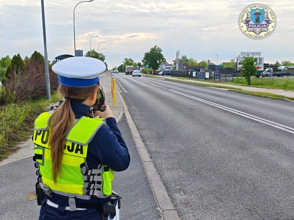 Policjantka stoi tyłem do zdjęcia, patrzy przez urządzenie do pomiaru prędkości jak jadą kierowcy samochodów.
