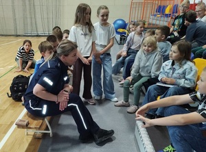W sali w szkole, policjantka siedzi na ławce, wokół niej są dzieci, rozmawiają z nią.