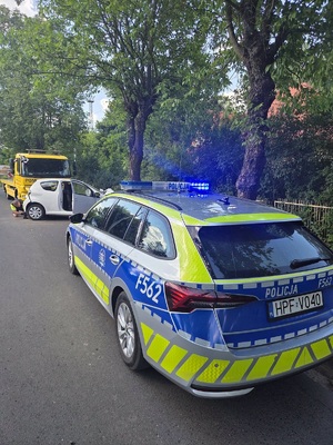 Radiowóz oznakowany z włączonymi światłami na miejscu wypadku, a tle pojazd który uderzył w drzewo.