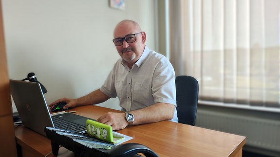 Za biurkiem siedzi mężczyzna w okularach, pozuje do zdjęcia.