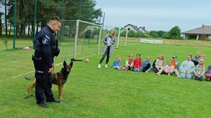 Pies i policjant są na boisku, koło nich siedzą dzieci. Policjant mówi do zebranych dzieci.