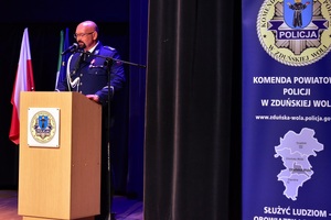 Na scenie, za mównicą jest policjant, mówi do zebranych.