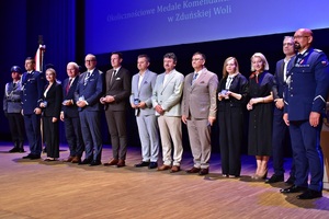 Na scenie stoi grupa osób. Otrzymali okolicznościowy medal. Pozują do zdjęcia.
