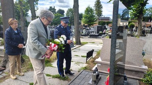 Policjant i mężczyzna podchodzą do tablicy na cmentarzu. Za nimi stoi kobieta.