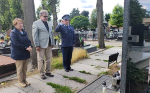 Przed tablicą na baczność stoi kobieta, mężczyzna i policjant, który oddaje honor.