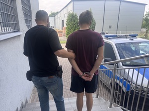 Nieumundurowany policjant prowadzący zatrzymanego mężczyznę w kajdankach do radiowozu.