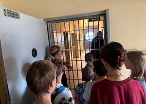Dzieci stoją przed drzwiami z kratą, patrzą na policjanta, który jest za kratami. Obok dzieci stoi policjantka.