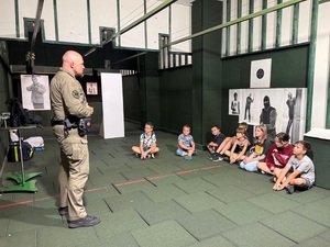 Pomieszczenie strzelnicy. Dzieci siedzą pod ścianą, a na wprost nich stoi policjant i mówi do nich.