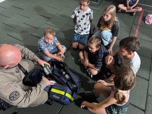 Policjant pokazuje dzieciom torbę w której są akcesoria niezbędne podczas udzielania pierwszej pomocy.