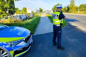 Policjant stoi tyłem do zdjęcia, patrzy przez wizjer urządzenia do pomiaru prędkości pojazdów.