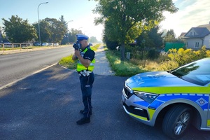 Policjant stoi przodem do zdjęcia, patrzy przez wizjer urządzenia do pomiaru prędkości pojazdów.