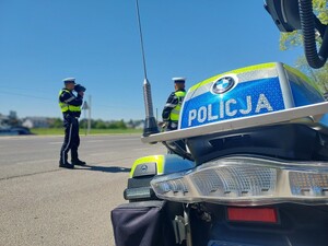 Przy drodze stoi motocykl służbowy oraz dwaj policjanci. Jeden z nich patrzy na drogę przez urządzenie do pomiaru prędkości.