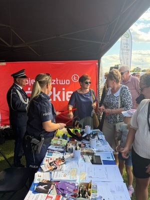 Zastępca Komendanta Powiatowego Policji w Zduńskiej Woli z policjantką, rozmawiający z seniorami przy stoisku profilaktycznym.