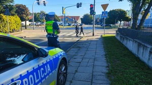 Policjant stoi obok radiowozu, patrzy na przejście dla pieszych, po którym przechodzą osoby.