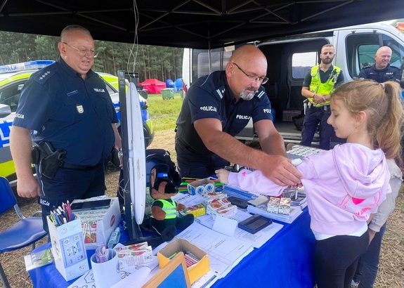 Pod namiotem, na stoisku profilaktycznym, policjanci rozmawiają z dziećmi, dają ulotki, odblaski i pieczątki.
