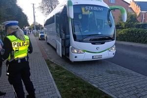 Policjantka stoi tyłem do zdjęcia. Autobus stoi na zatoczce. W tle radiowóz i chodnik po którym idzie młodzież.