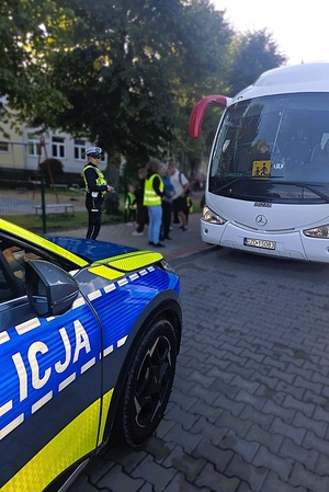 Radiowóz, za nim stoi policjantka. W tle autobus i dzieci wsiadające do niego.
