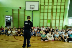 Sala gimnastyczna. Tyłem do zdjęcia stoi policjantka, przed nią na podłodze siedzą dzieci, słuchają co ona do nich mówi.