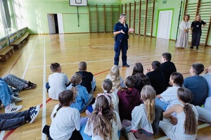 Sala gimnastyczna. Stoi policjantka, przed nią na podłodze siedzą dzieci, słuchają co ona do nich mówi.