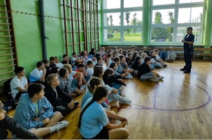 Sala gimnastyczna. Policjantka stoi, przed nią na podłodze siedzą dzieci, słuchają co ona do nich mówi.